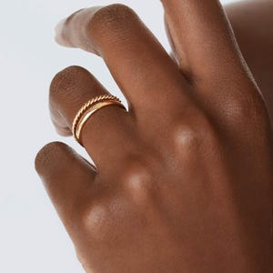 May include: Two gold rings on a finger. The top ring is a twisted band and the bottom ring is a simple band.