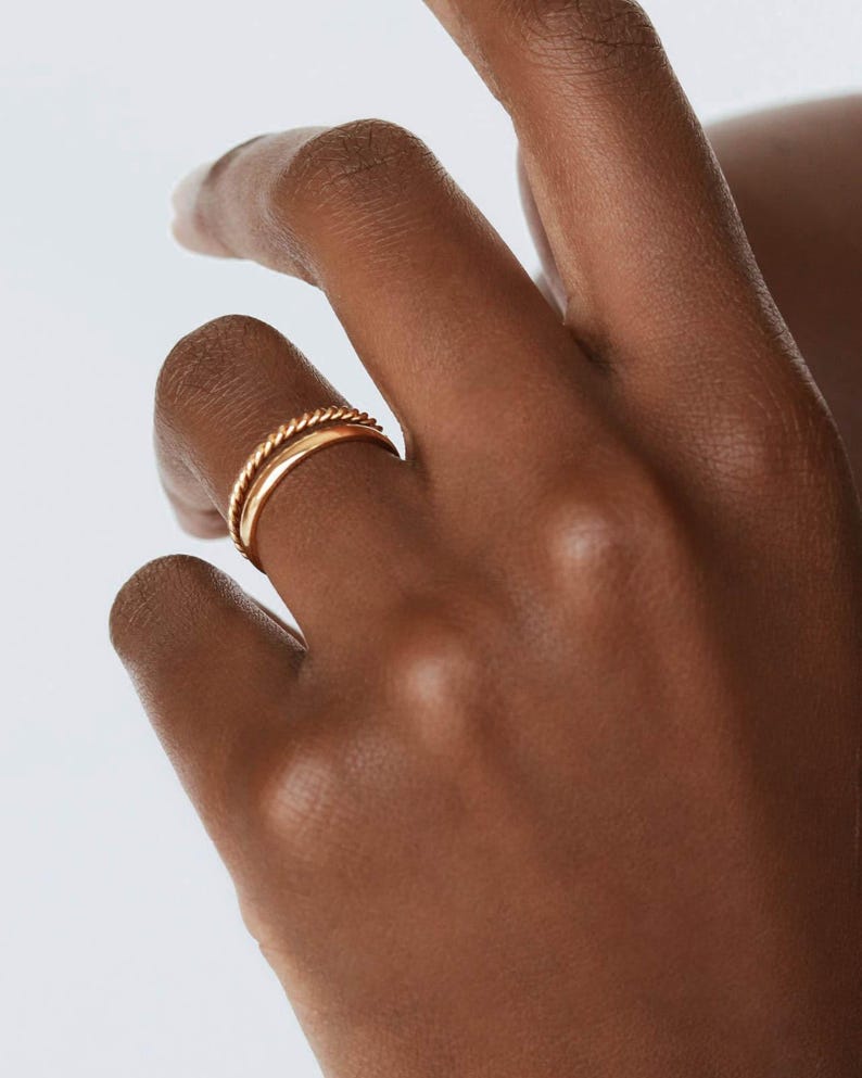 May include: Two gold rings on a finger. The top ring is a twisted band and the bottom ring is a simple band.