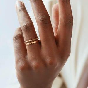 May include: Two gold rings on a finger. The top ring is a twisted band and the bottom ring is a simple band.