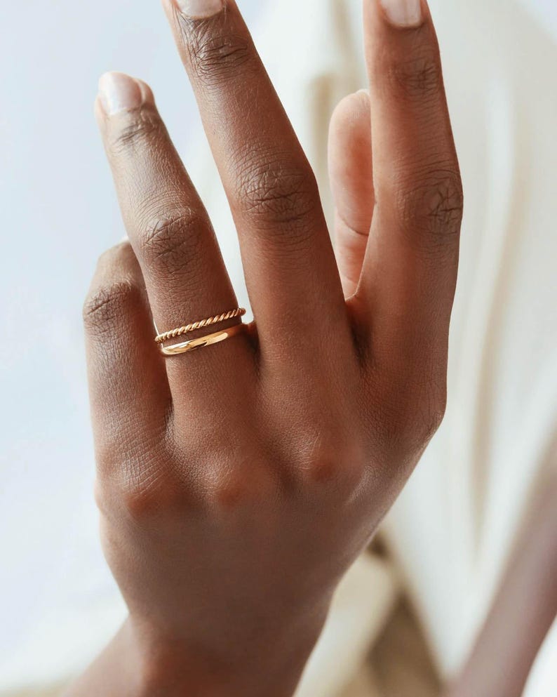May include: Two gold rings on a finger. The top ring is a twisted band and the bottom ring is a simple band.