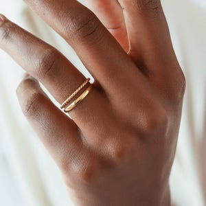 May include: Two gold rings on a person's finger. The top ring is a twisted band and the bottom ring is a simple band.