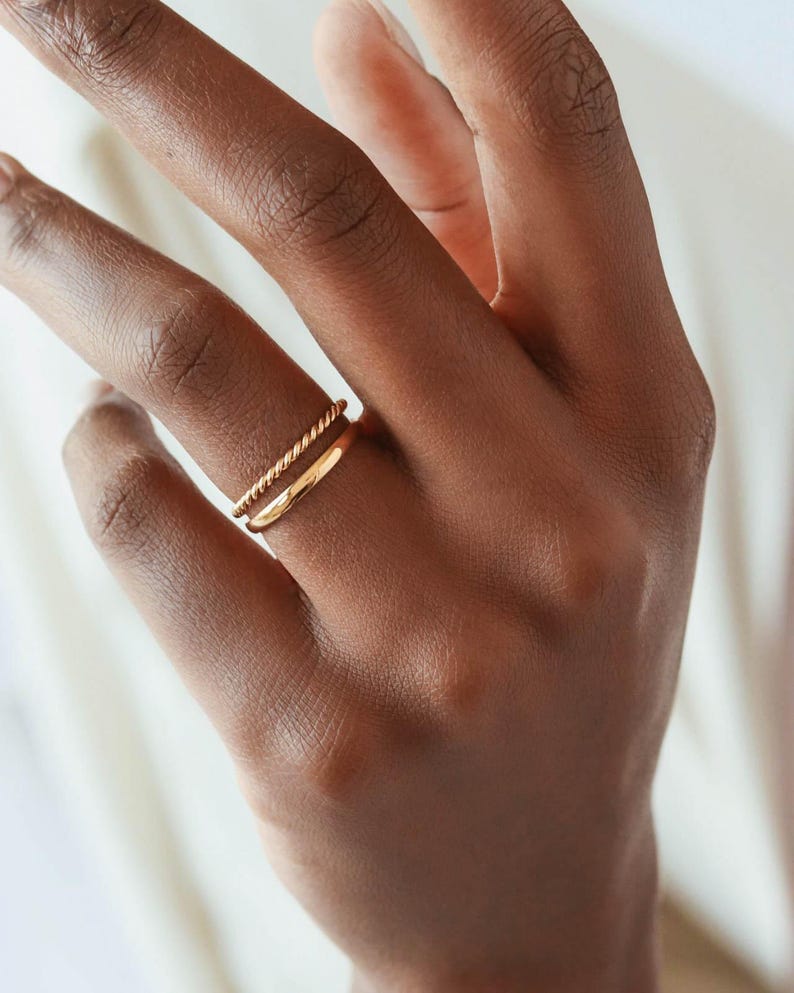 May include: Two gold rings on a person's finger. The top ring is a twisted band and the bottom ring is a simple band.