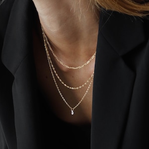 Op de afbeelding: Een gelaagde gouden ketting met een klein traanvormig hangertje. De ketting wordt gedragen door iemand met een zwart blazer.