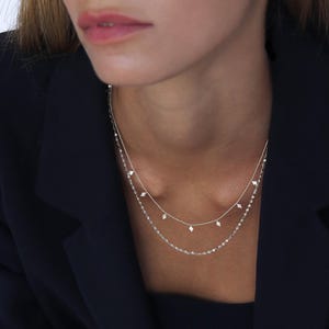 Op de afbeelding: Een zilveren ketting met een delicate ketting en kleine diamantvormige bedels die eraan hangen. De ketting is gelegd over een dikkere zilveren ketting.