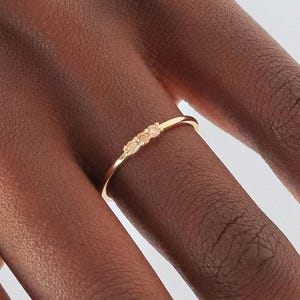 Op de afbeelding: Een delicate gouden ring met drie kleine, ronde, lichtbruine edelstenen. De ring heeft een minimalistisch ontwerp en een gepolijste afwerking. De ring wordt aan een vinger gedragen, wat de eenvoudige elegantie benadrukt. De edelstenen zijn in een rij geplaatst.