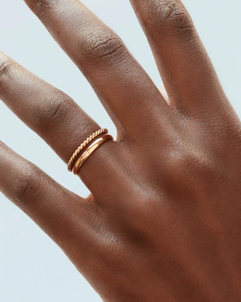 May include: Two gold rings on a finger. The top ring is a twisted band and the bottom ring is a simple band.