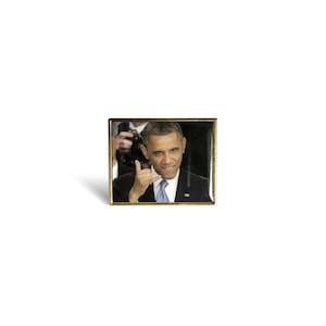 Peut inclure: Une épingle dorée avec une photo de Barack Obama faisant le geste 'appelle-moi'. Il porte un costume sombre et une cravate bleue.