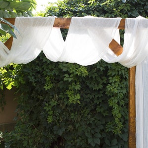 May include: A wooden wedding archway decorated with white sheer fabric draped over the top.