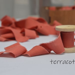 May include: A spool of terracotta colored fabric ribbon with a wooden spool. The ribbon is frayed and has a rustic look.