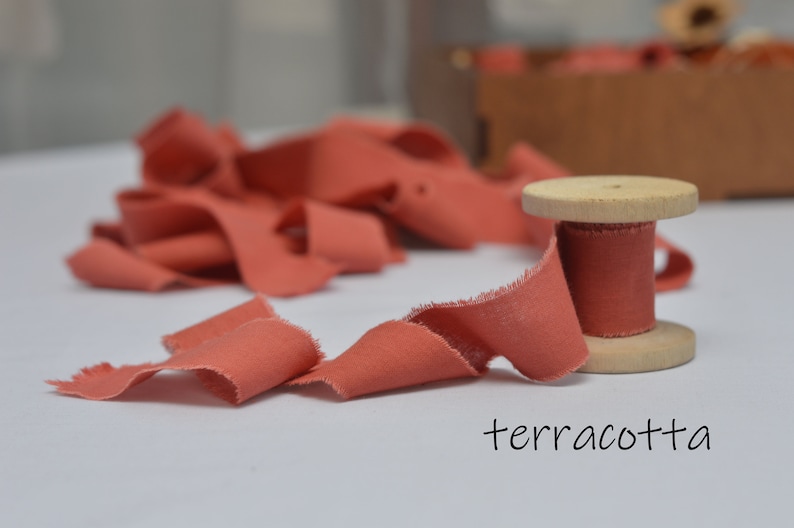 May include: A spool of terracotta colored fabric ribbon with a wooden spool. The ribbon is frayed and has a rustic look.