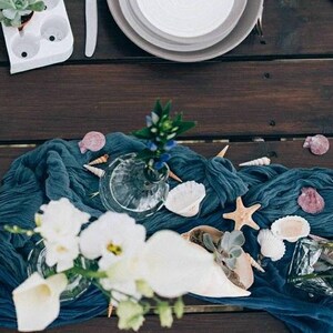 Può includere: Un runner da tavolo in lino blu con fiori bianchi, conchiglie e una stella marina. Il runner è su un tavolo in legno scuro.