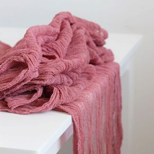 May include: Peach pink fabric table runner with a textured, crinkled look. The runner is draped over a white table.