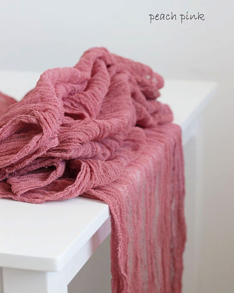 May include: Peach pink fabric table runner with a textured, crinkled look. The runner is draped over a white table.