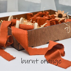 May include: A wooden box filled with burnt orange fabric ribbon spools. The ribbon is frayed and has a rustic look. The box is filled with shredded paper and a small flower.