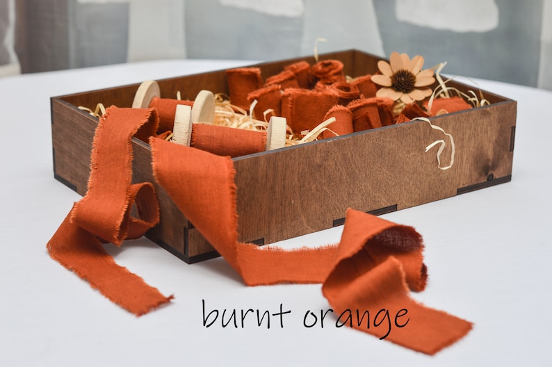 May include: A wooden box filled with burnt orange fabric ribbon spools. The ribbon is frayed and has a rustic look. The box is filled with shredded paper and a small flower.