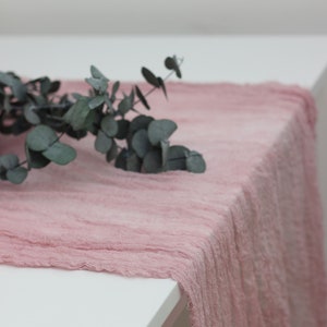 May include: Blush pink table runner made of a soft, textured fabric. The runner is draped over a white table with a sprig of eucalyptus leaves on top.