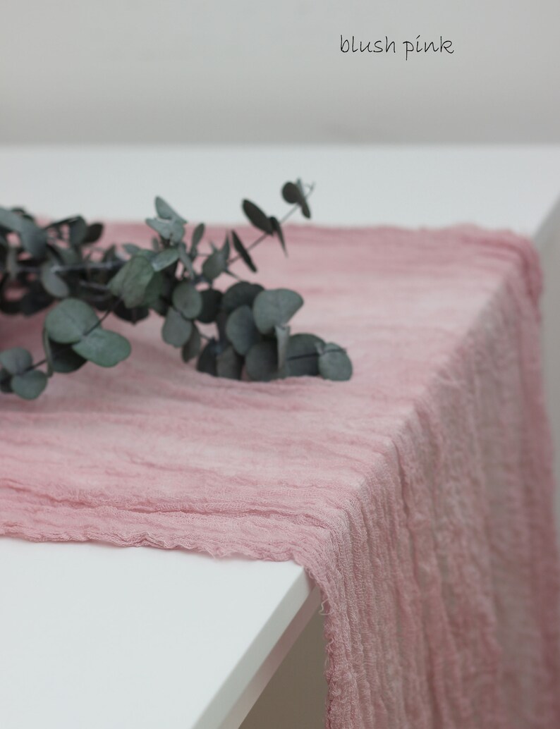 May include: Blush pink table runner made of a soft, textured fabric. The runner is draped over a white table with a sprig of eucalyptus leaves on top.