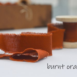 May include: A spool of burnt orange fabric ribbon with frayed edges. The ribbon is wound around a wooden spool.