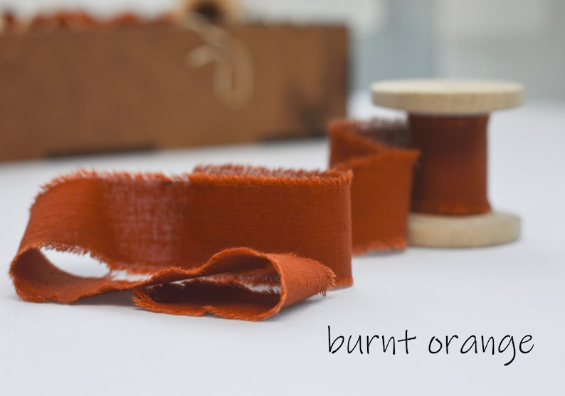 May include: A spool of burnt orange fabric ribbon with frayed edges. The ribbon is wound around a wooden spool.