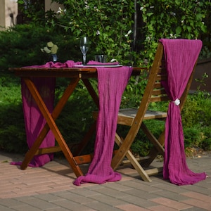 May include: A wooden table with a pink table runner and a pink chair sash. The table is set with a glass, a plate, and a napkin. The table and chair are outdoors on a brick patio.