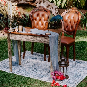 May include: A rustic wooden table with a distressed white finish, set with two brown leather chairs, a blue cloth runner, and a decorative feather pen. The table is surrounded by green grass and a blue and white patterned rug.