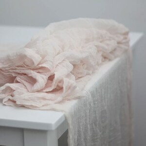 May include: A soft, blush-colored table runner made of sheer fabric. The runner is draped over a white table and has a delicate, ruffled texture.