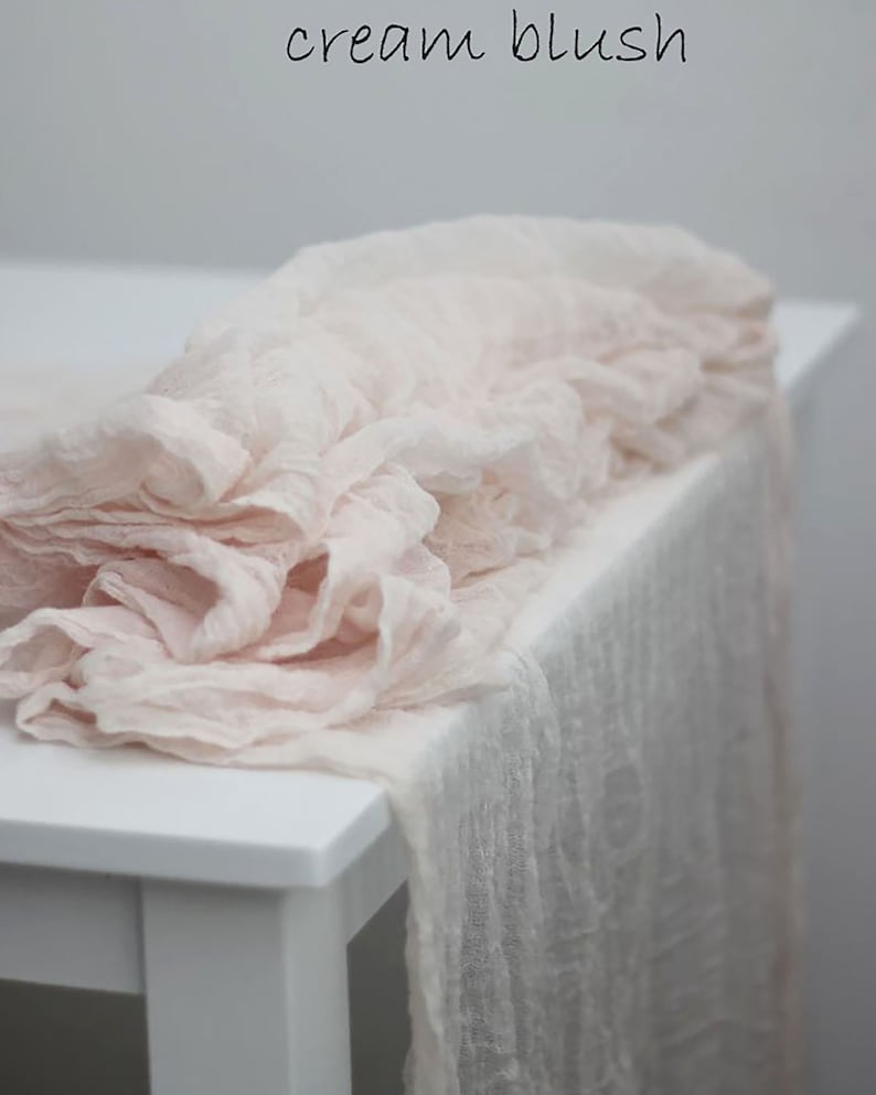 May include: A soft, blush-colored table runner made of sheer fabric. The runner is draped over a white table and has a delicate, ruffled texture.