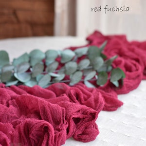 May include: A red fuchsia fabric runner with a green eucalyptus garland on top. The runner is made of a soft, crinkled fabric and has a delicate, romantic feel.