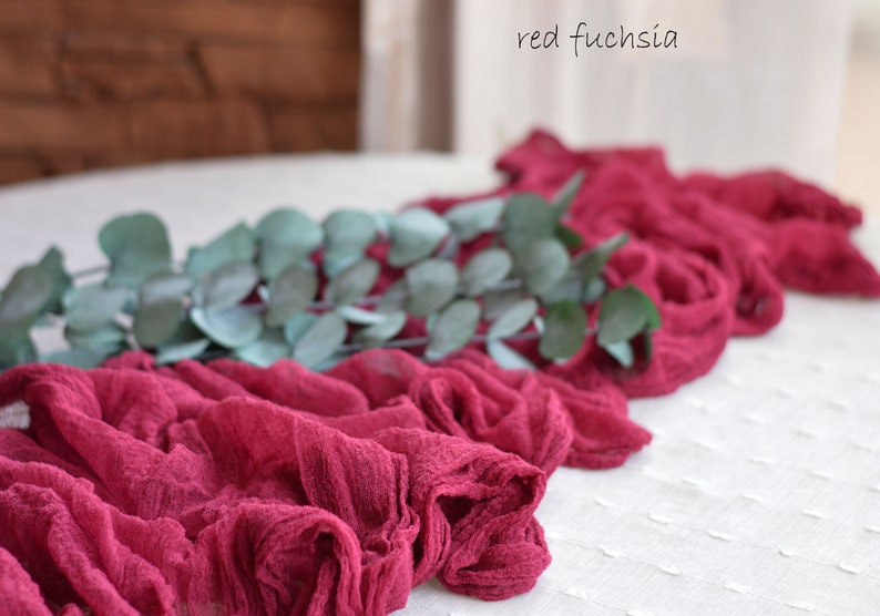 May include: A red fuchsia fabric runner with a green eucalyptus garland on top. The runner is made of a soft, crinkled fabric and has a delicate, romantic feel.