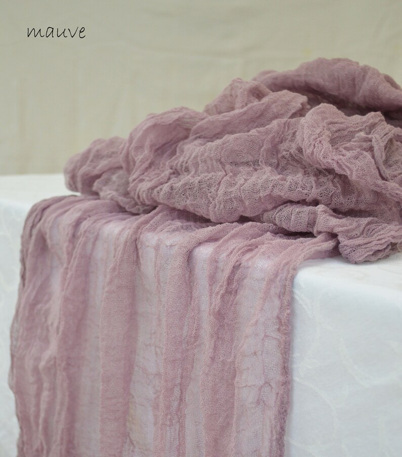 May include: Mauve colored, textured, sheer fabric table runner. The fabric is loosely woven and has a crinkled appearance.