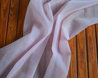 May include: A pale pink sheer fabric draped over a wooden surface. The fabric is smooth and delicate, with a soft, flowing texture.