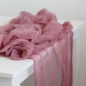 May include: Dusty rose colored sheer fabric table runner. The fabric is gathered and draped over a white table.