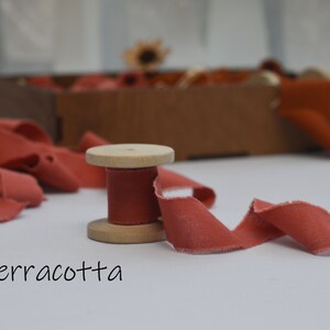 May include: A spool of terracotta colored fabric ribbon on a wooden spool. The ribbon is unraveled and laying on a white surface.