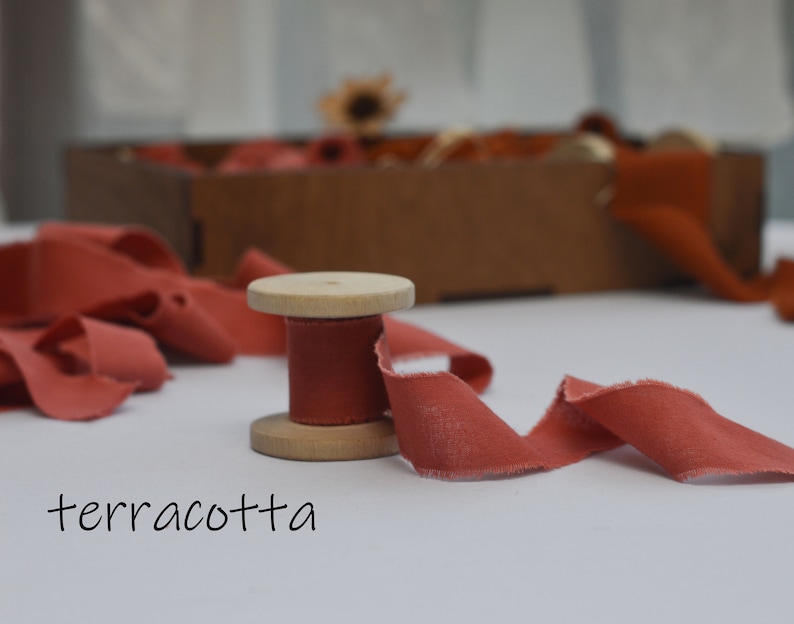 May include: A spool of terracotta colored fabric ribbon on a wooden spool. The ribbon is unraveled and laying on a white surface.