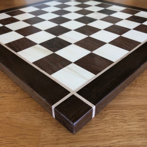 May include: A wooden chessboard with a dark brown and light wood checkered pattern. The board has a dark brown border with a light wood inlay.