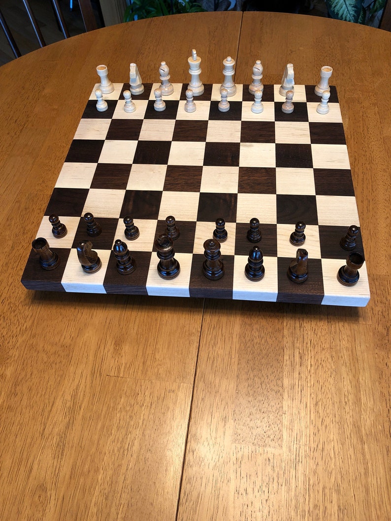 May include: A wooden chessboard with black and white squares. The chess pieces are arranged in their starting positions. The white pieces are made of light wood and the black pieces are made of dark wood.