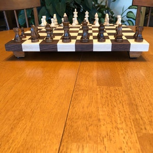 May include: A wooden chessboard with black and white squares. The chess pieces are made of wood and are in the starting position. The chessboard is on a wooden table.