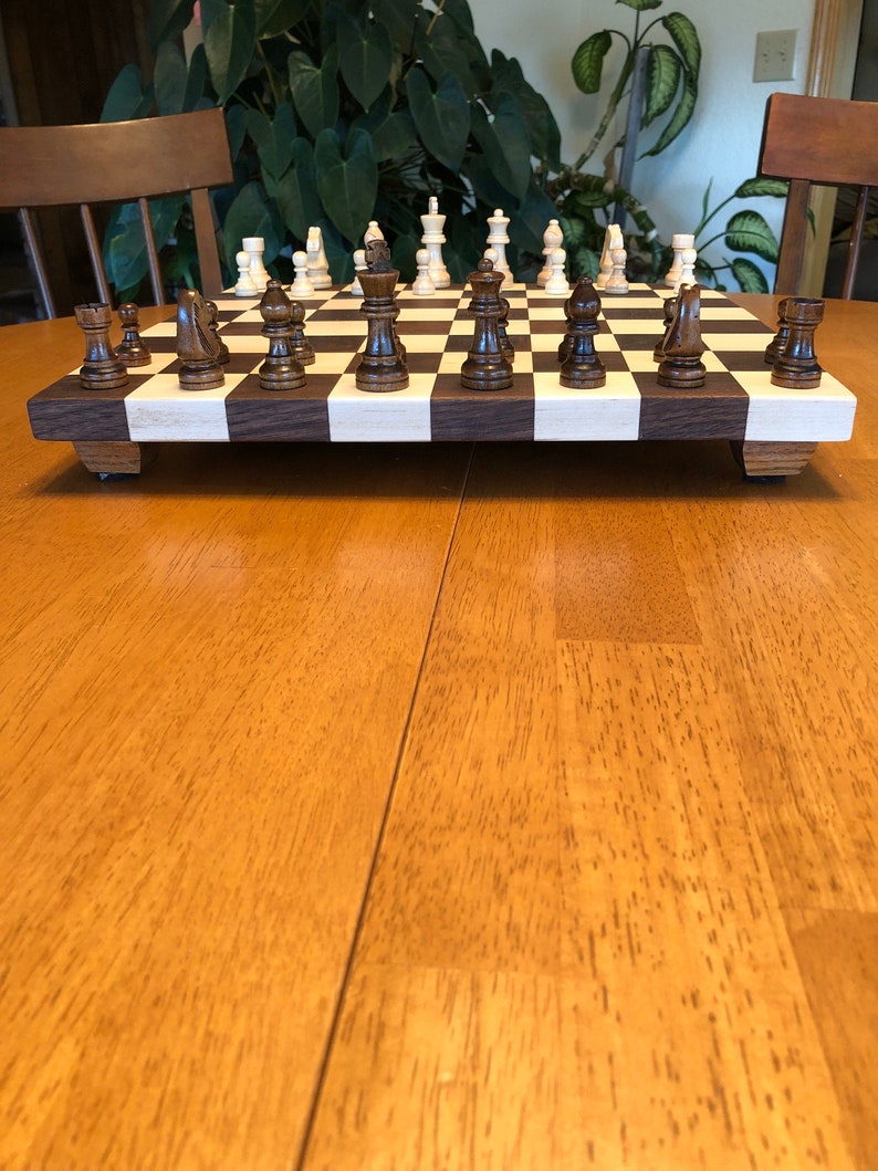 May include: A wooden chessboard with black and white squares. The chess pieces are made of wood and are in the starting position. The chessboard is on a wooden table.
