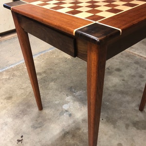 May include: A wooden chess table with a checkerboard inlay top and a drawer. The table has four legs and is made of dark wood.