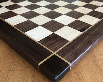 Puede incluir: Un tablero de ajedrez de madera con un borde de nogal oscuro y una superficie de juego de arce claro. El tablero tiene una incrustación de madera clara alrededor del borde.