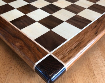 May include: A wooden chessboard with a dark brown and light wood checkered pattern. The board has a dark brown wood frame with a white inlay around the edge.