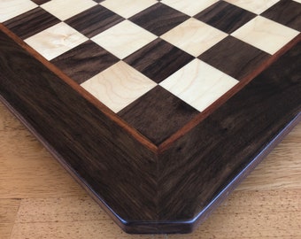 Puede incluir: Un tablero de ajedrez de madera con un marco marrón oscuro y una superficie de juego de madera clara. El tablero está hecho de madera de nogal y arce.