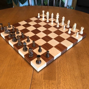 May include: A wooden chessboard with a light brown and dark brown checkered pattern. The board is set up with white and black chess pieces. The pieces are arranged in a standard starting position for a game of chess.