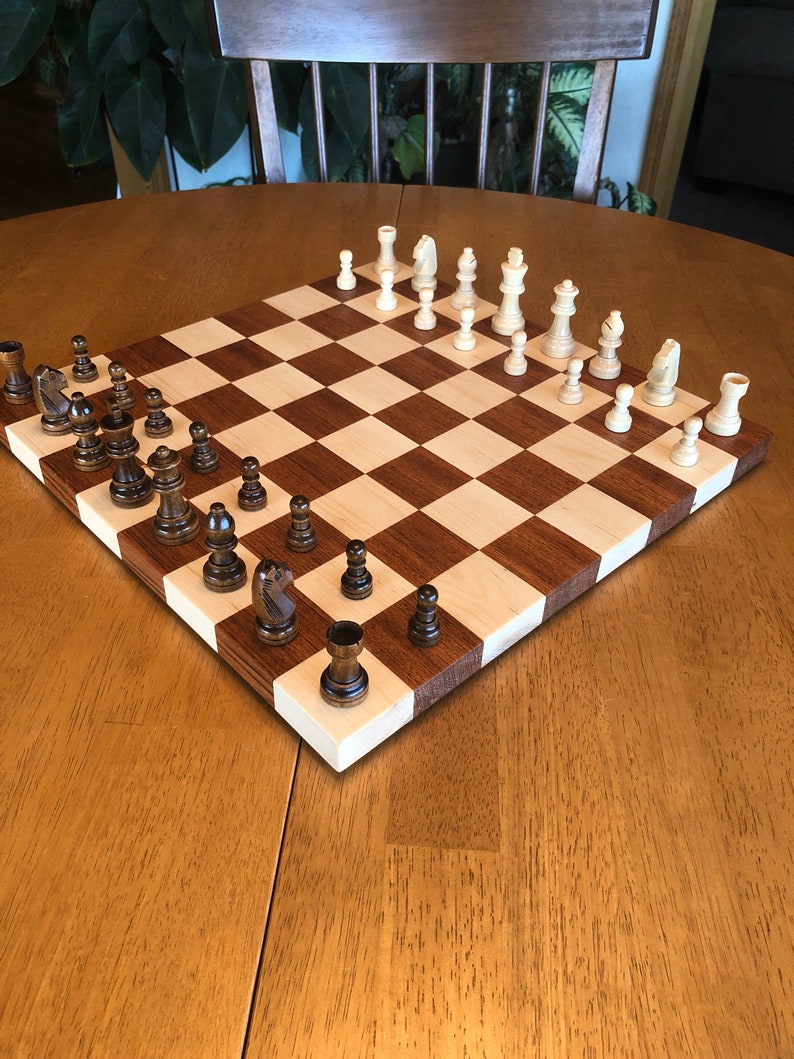 May include: A wooden chessboard with a light brown and dark brown checkered pattern. The board is set up with white and black chess pieces. The pieces are arranged in a standard starting position for a game of chess.