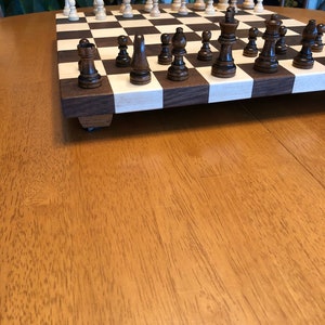 May include: A wooden chessboard with black and white squares. The board is set up for a game of chess with black and white pieces in place. The board is on a wooden table.