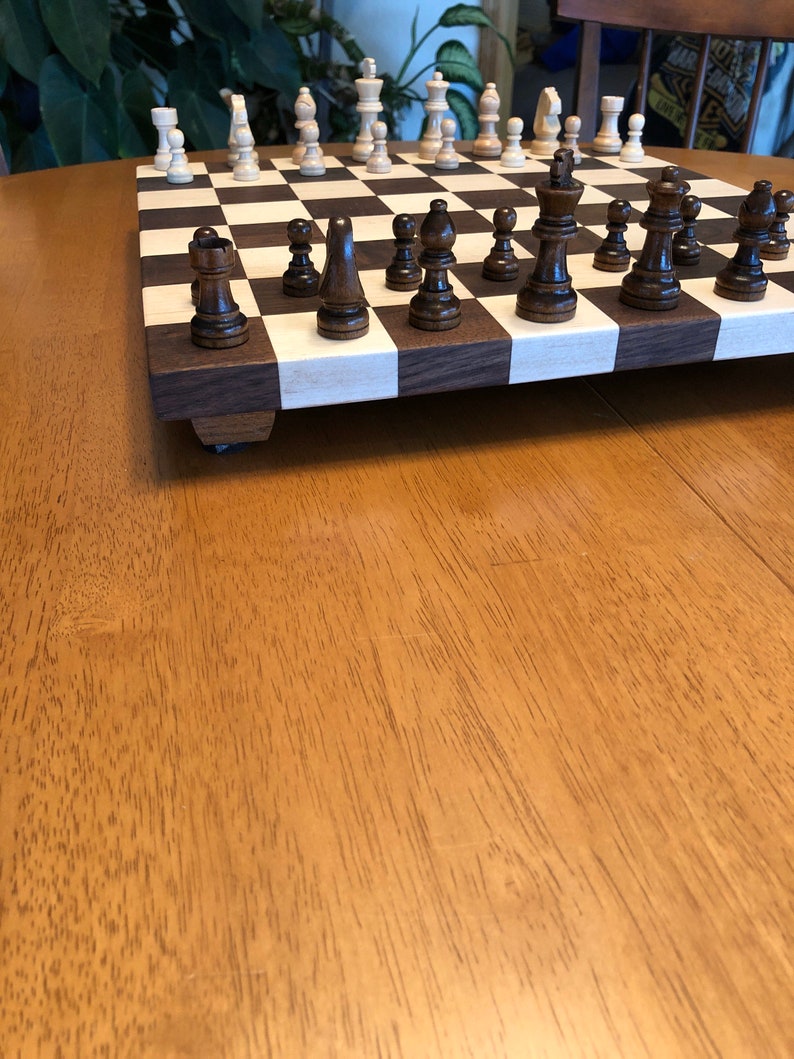 May include: A wooden chessboard with black and white squares. The board is set up for a game of chess with black and white pieces in place. The board is on a wooden table.