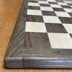May include: A wooden chessboard with a dark brown border and alternating light and dark squares. The board is made of wood and has a polished finish. The chessboard is designed for playing chess.