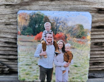May include: A family of four poses for a photo in a field. The father is wearing a white shirt and jeans, and he is holding a young boy on his shoulders. The mother is wearing a white sweater and jeans, and she is holding a young girl in her arms. The children are both wearing casual clothing. The background is a field with trees and fall foliage.