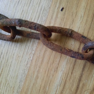 May include: Close-up of a rusty metal chain with large, oval links. The chain shows signs of age and wear, with a reddish-brown color and areas of corrosion. The chain is lying on a light brown wooden surface.