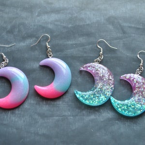 May include: A pair of crescent moon shaped earrings with glitter and ombre designs. One pair is pink and blue, the other is teal and purple.